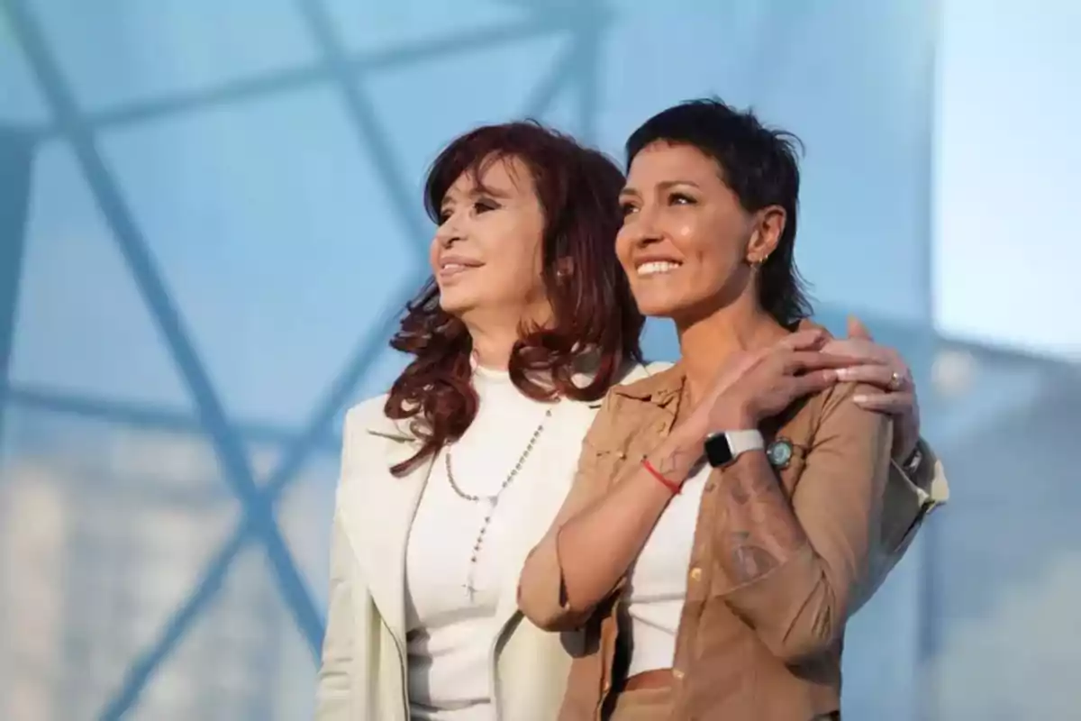 Dos mujeres sonríen y miran hacia el horizonte mientras se abrazan, con un fondo de estructura moderna y cielo azul.