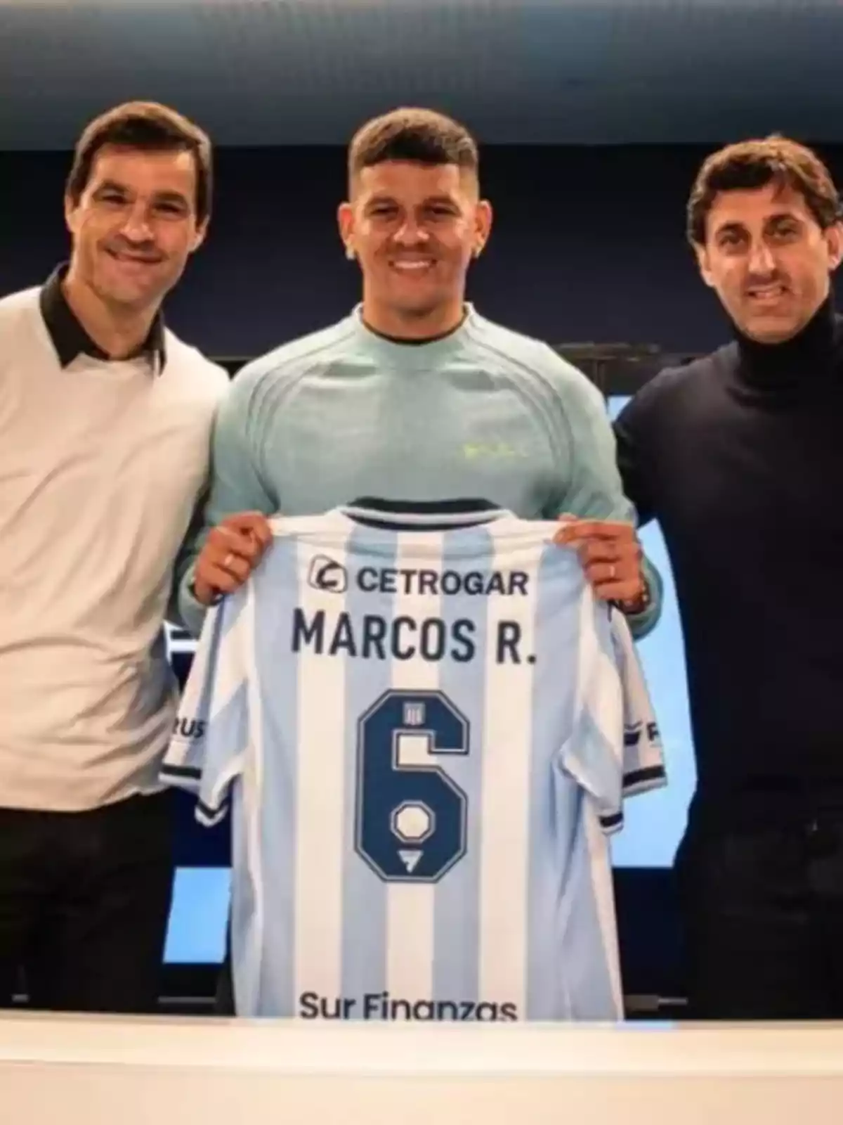 Tres hombres sonríen mientras el del centro sostiene una camiseta de fútbol celeste y blanca con el nombre Marcos R. y el número 6.