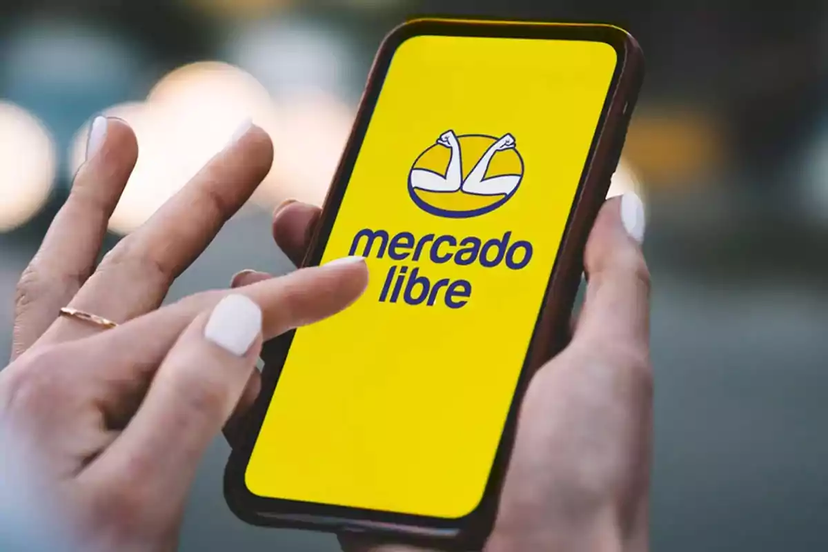 Persona usando un teléfono móvil que muestra la pantalla de inicio de la aplicación Mercado Libre con fondo amarillo y el logo visible
