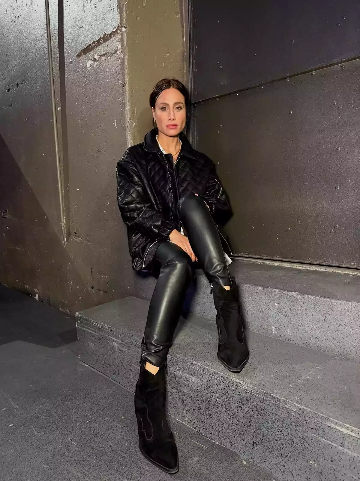 Woman sitting on concrete stairs wearing a black puffer jacket, leather pants, and black boots, with a serious expression and an industrial wall background Woman sitting on concrete stairs wearing a black puffer jacket, leather pants, and black boots, with a serious expression and an industrial wall background