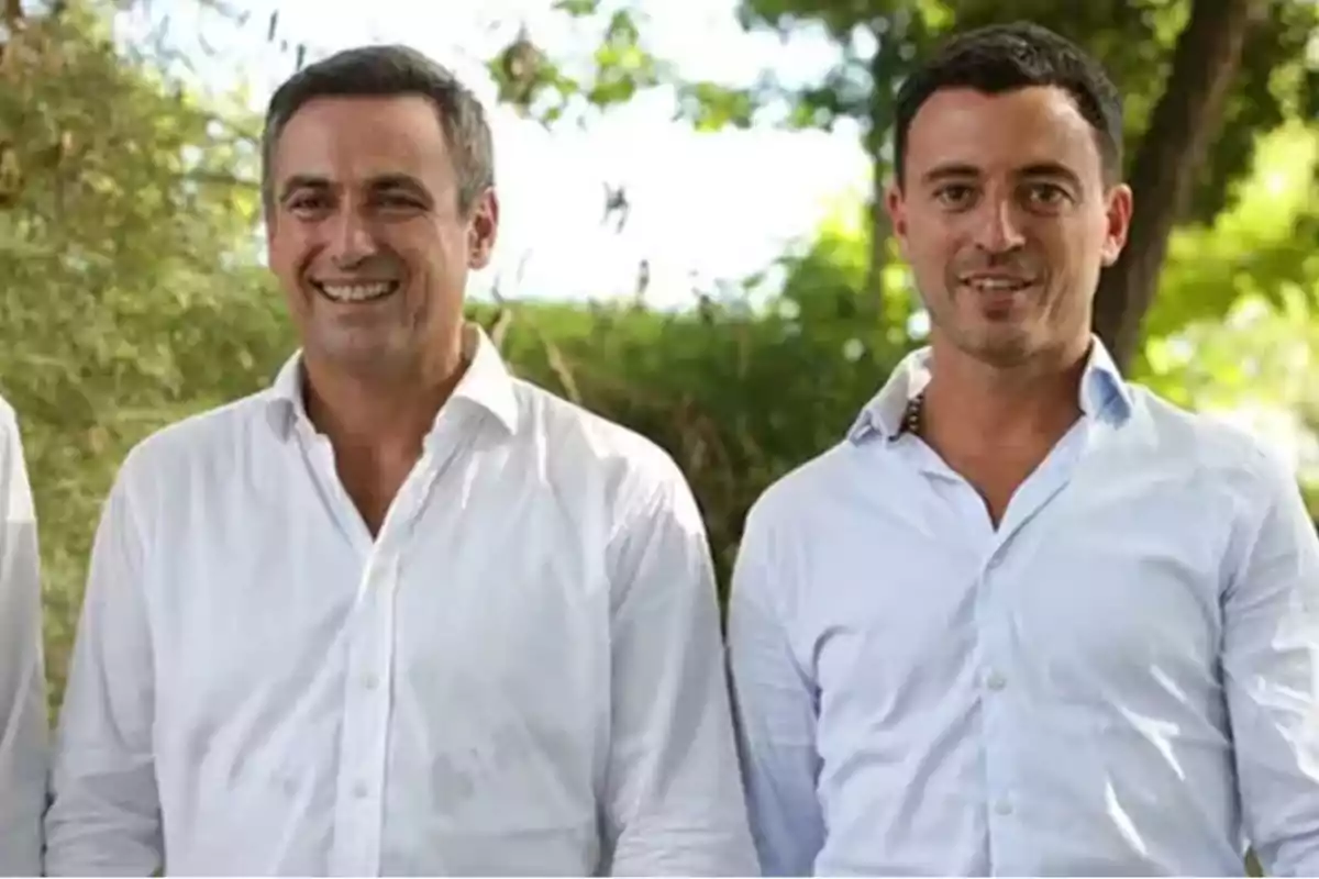 Dos hombres sonrientes vestidos con camisas blancas posan al aire libre con árboles de fondo