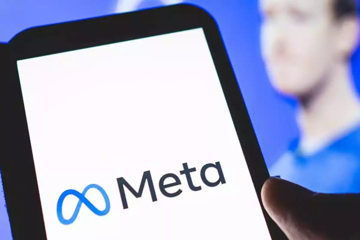 Persona sosteniendo un dispositivo móvil que muestra el logo y el nombre de Meta en la pantalla, con una figura desenfocada al fondo.