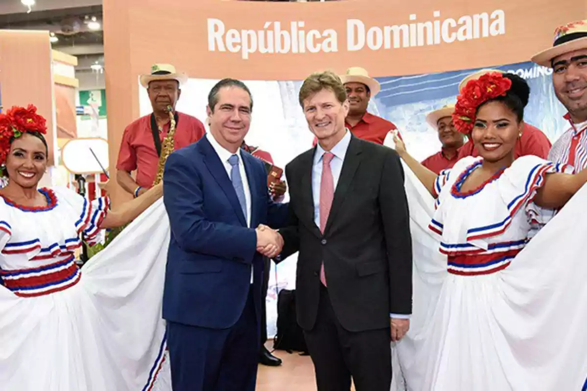 Dos hombres de traje se dan la mano frente a un grupo de personas con trajes típicos dominicanos y un letrero que dice República Dominicana Dos hombres de traje se dan la mano frente a un grupo de personas con trajes típicos dominicanos y un letrero que dice República Dominicana