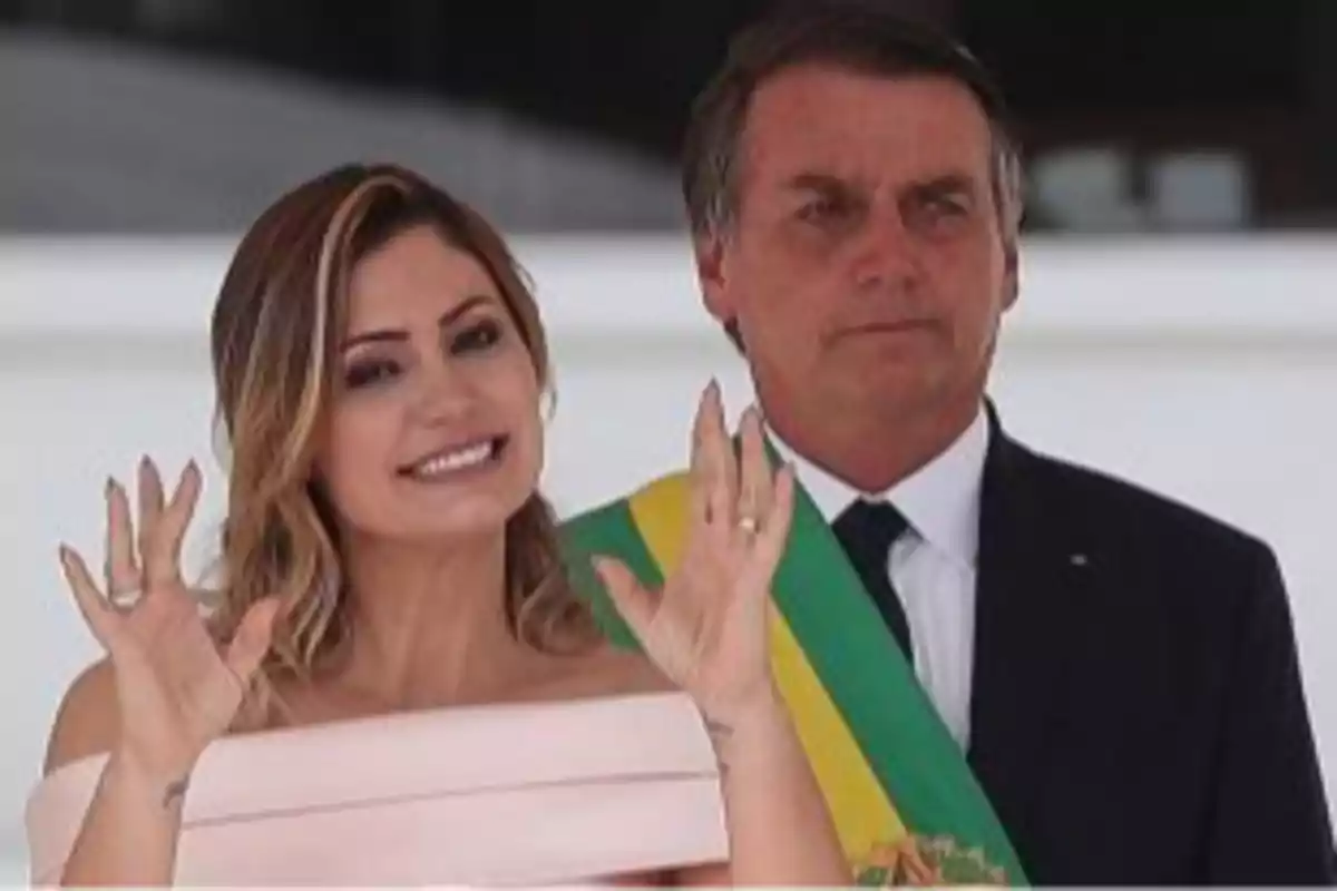 A smiling woman waves with both hands raised while a man wearing a green and yellow presidential sash stands beside her.