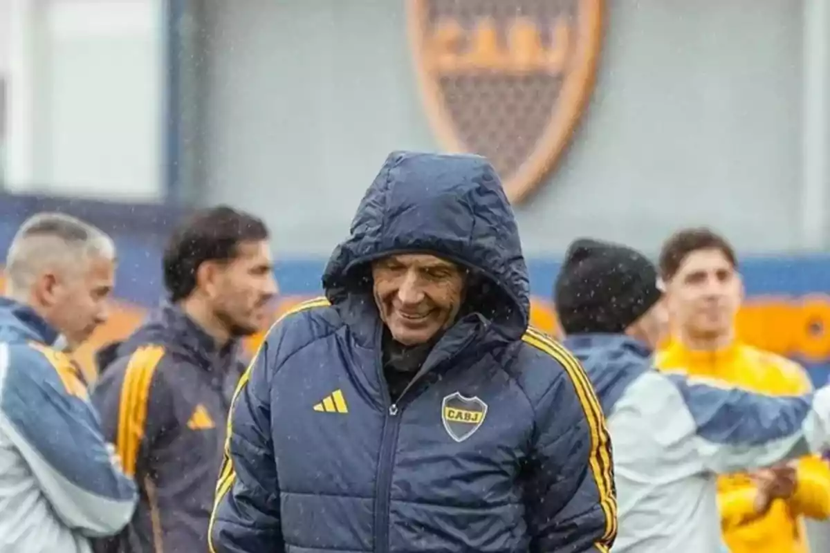 Entrenador de fútbol sonriente con campera azul y capucha durante un entrenamiento bajo la lluvia junto a su equipo Entrenador de fútbol sonriente con campera azul y capucha durante un entrenamiento bajo la lluvia junto a su equipo