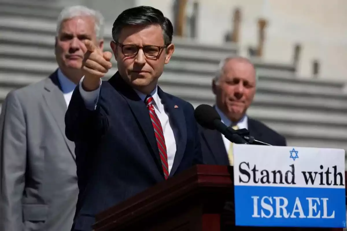 A man wearing glasses and a dark suit points forward while speaking at a podium with a sign that says "Stand with Israel," and two men in suits stand behind him on some stairs.