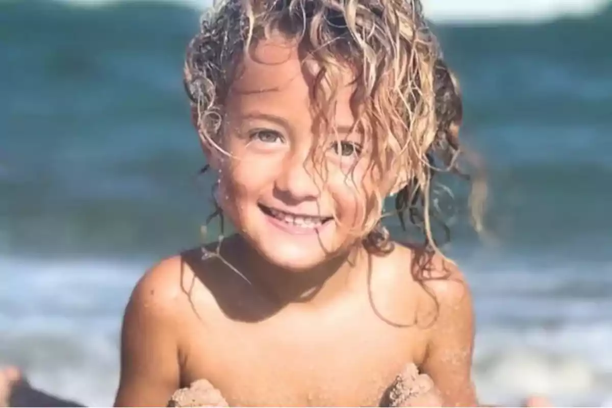Niña sonriente con el cabello rizado y mojado jugando en la playa con el mar de fondo Niña sonriente con el cabello rizado y mojado jugando en la playa con el mar de fondo