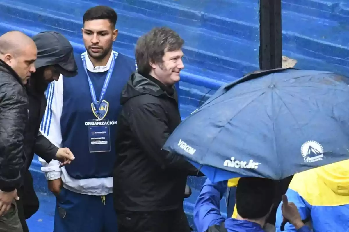 Several people wearing dark clothing and holding a blue umbrella are in a stadium with seats of the same color. Several people wearing dark clothing and holding a blue umbrella are in a stadium with seats of the same color.