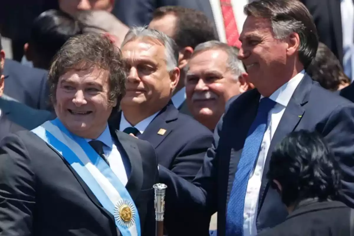 A man wearing a light blue and white presidential sash smiles as he is congratulated by another man in a blue suit at a public event surrounded by people. A man wearing a light blue and white presidential sash smiles as he is congratulated by another man in a blue suit at a public event surrounded by people.