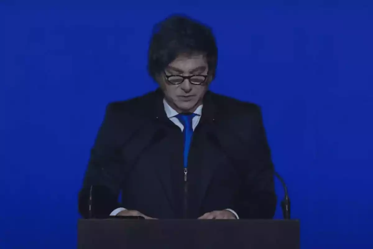 Dark-haired man with glasses reading a speech at a podium in front of a blue background