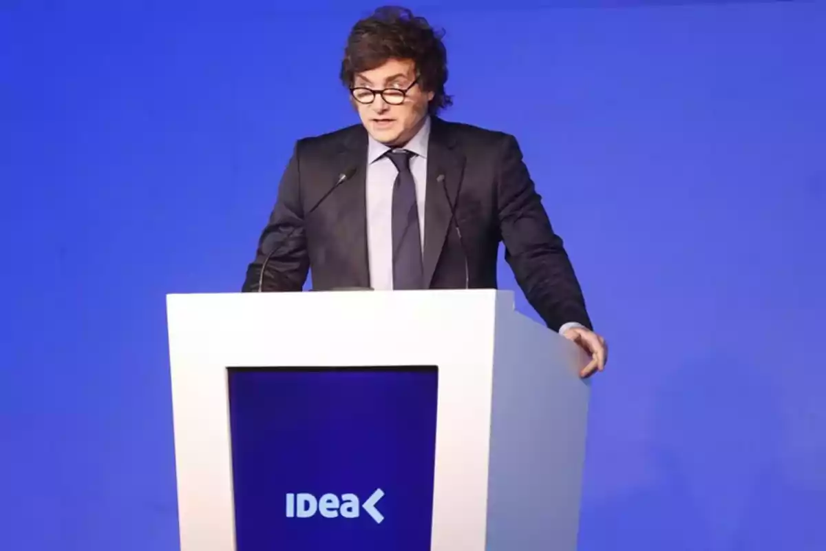 Hombre de cabello castaño y gafas hablando en un atril blanco con el logo de IDEA sobre un fondo azul Hombre de cabello castaño y gafas hablando en un atril blanco con el logo de IDEA sobre un fondo azul