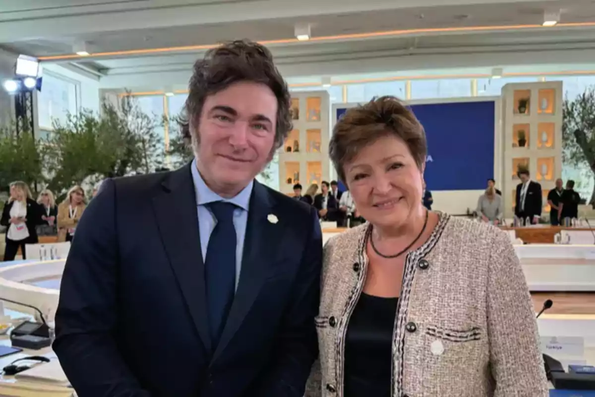 Dos personas sonríen y posan juntas en un evento formal con varias personas y mesas al fondo en un ambiente iluminado y moderno