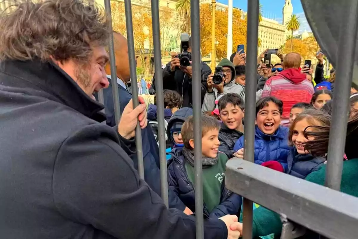 Un hombre sonriente saluda a un grupo de niños felices a través de una reja mientras varias personas toman fotos en el fondo