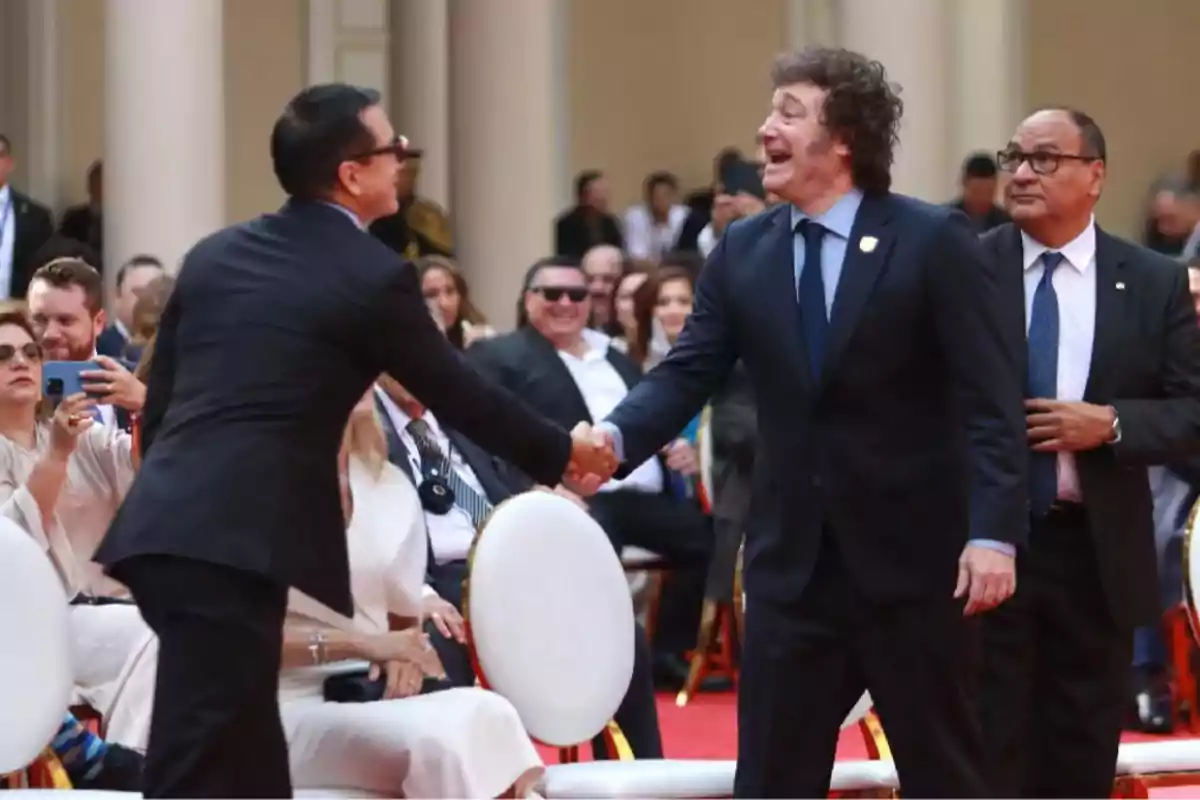 Dos hombres de traje se saludan con un apretón de manos en un evento formal mientras varias personas observan y sonríen en el fondo