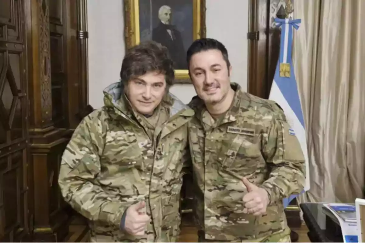 Two men dressed in camouflage clothing pose together in an office with an Argentine flag and a portrait on the wall in the background.