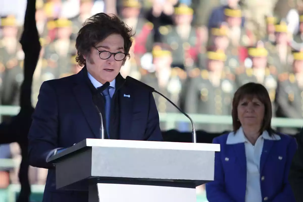 Un hombre con gafas y traje oscuro habla en un podio mientras una mujer con saco azul lo observa y varias personas con uniforme militar están desenfocadas al fondo Un hombre con gafas y traje oscuro habla en un podio mientras una mujer con saco azul lo observa y varias personas con uniforme militar están desenfocadas al fondo