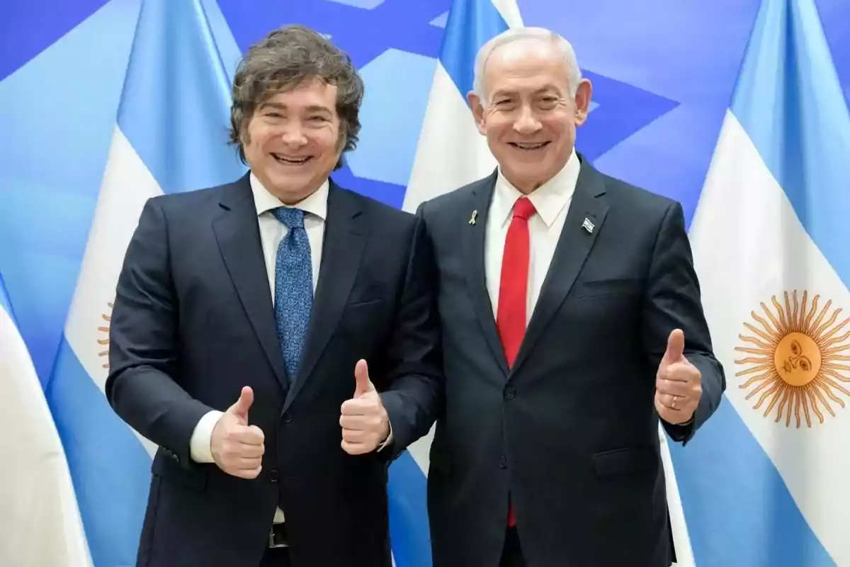 Dos hombres de traje sonríen y posan juntos haciendo el gesto de pulgar arriba frente a banderas de Argentina e Israel