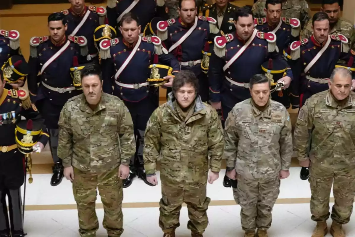 A group of military personnel in camouflage uniforms and soldiers in dress uniforms pose in formation inside a building.