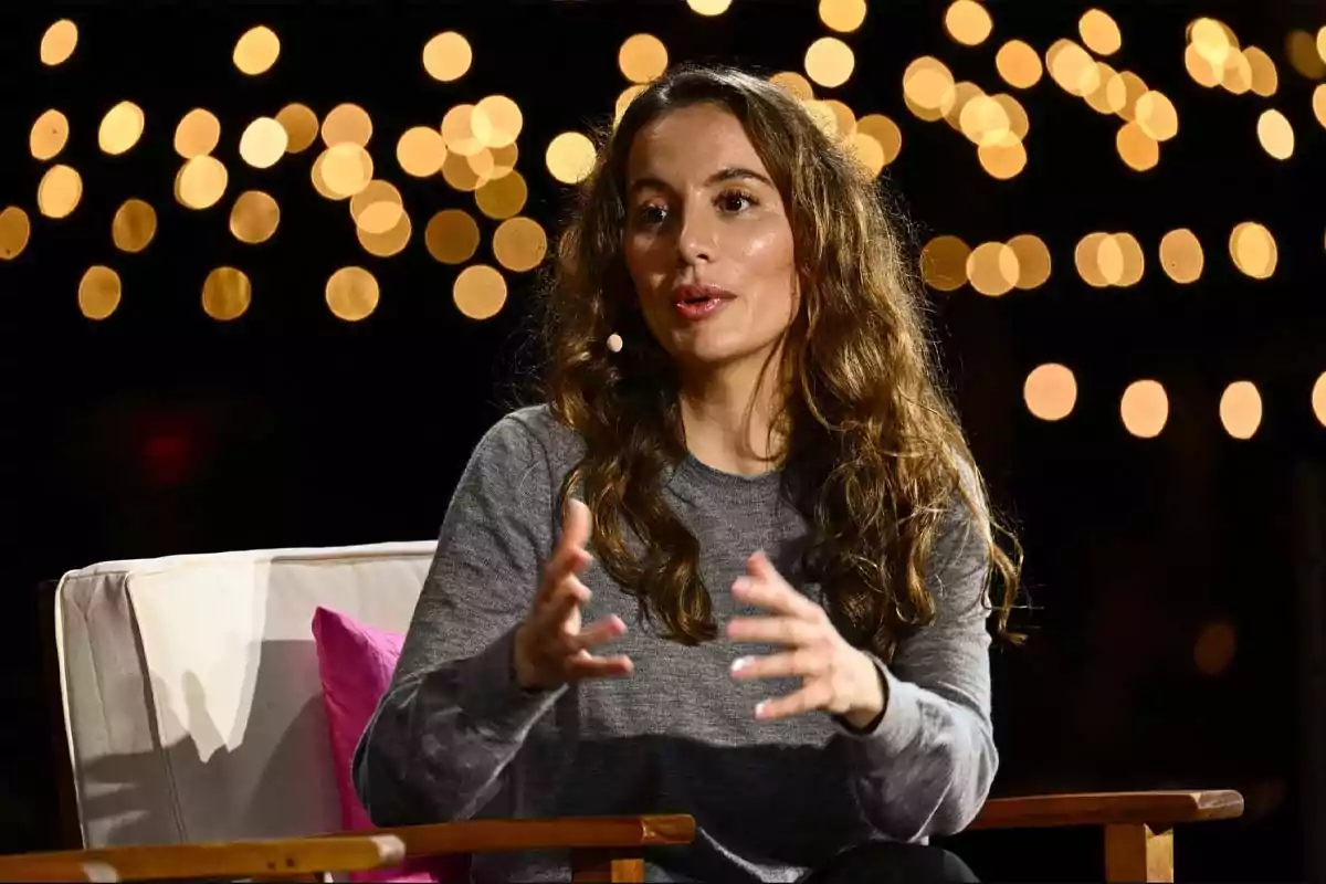 Mujer de cabello largo y ondulado hablando sentada en un sillón con luces desenfocadas de fondo