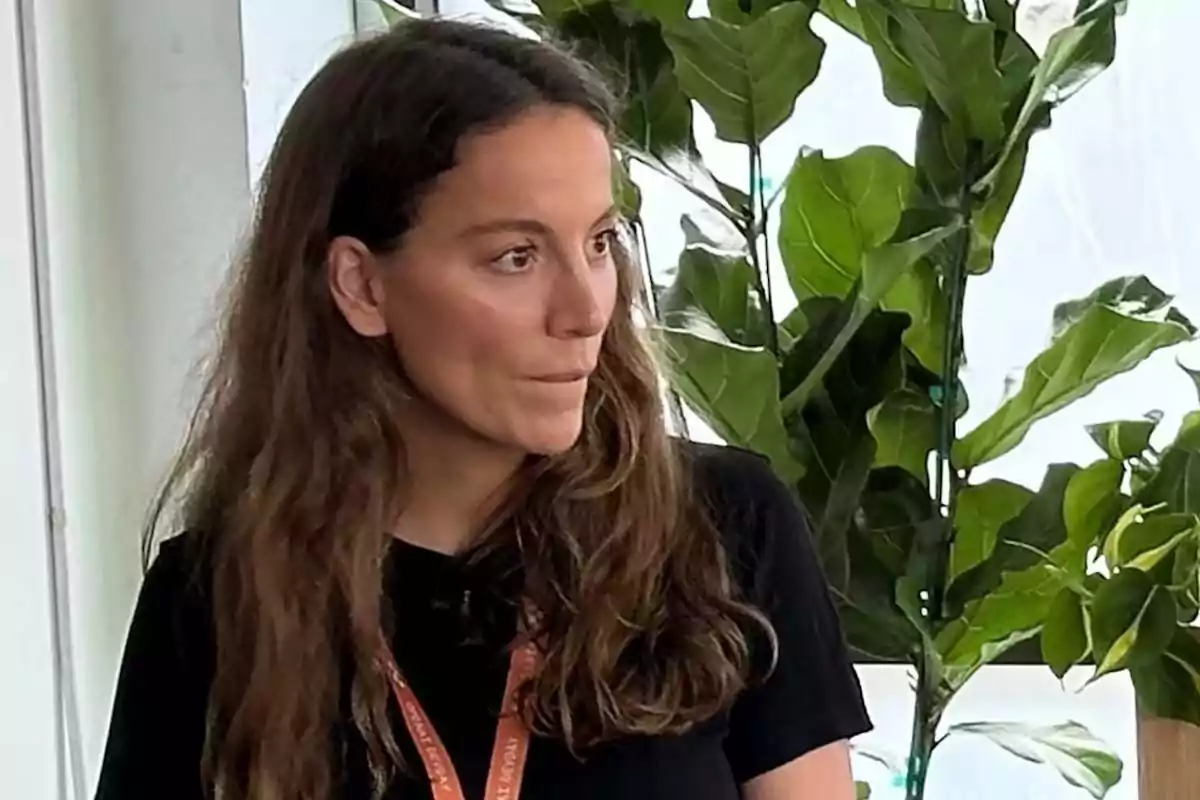 Mujer de cabello largo y castaño con camiseta negra y gafete naranja, de pie frente a plantas verdes