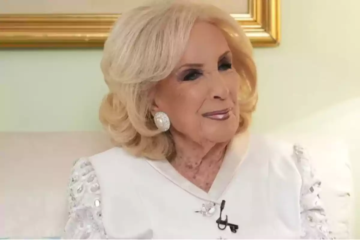 Mujer mayor de cabello rubio y corto, vestida elegantemente de blanco, sentada y sonriendo suavemente Mujer mayor de cabello rubio y corto, vestida elegantemente de blanco, sentada y sonriendo suavemente