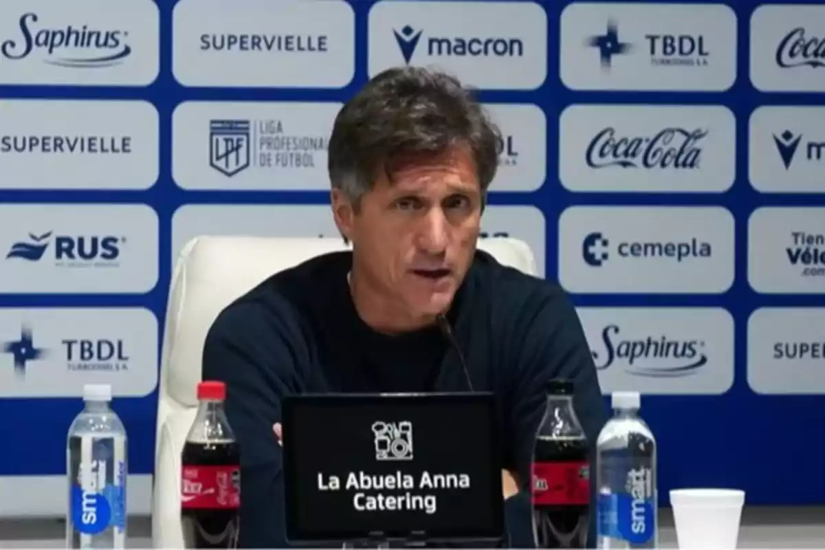Brown-haired man sitting at a press conference with a sponsor backdrop and bottles of water and soda on the table Brown-haired man sitting at a press conference with a sponsor backdrop and bottles of water and soda on the table
