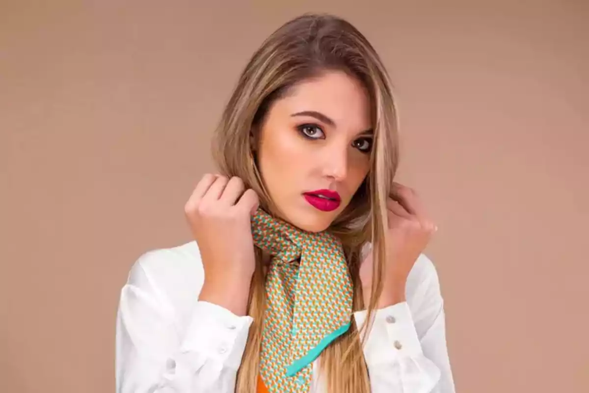 Woman with long blonde hair wearing a white jersey and a colorful scarf around her neck against a beige background