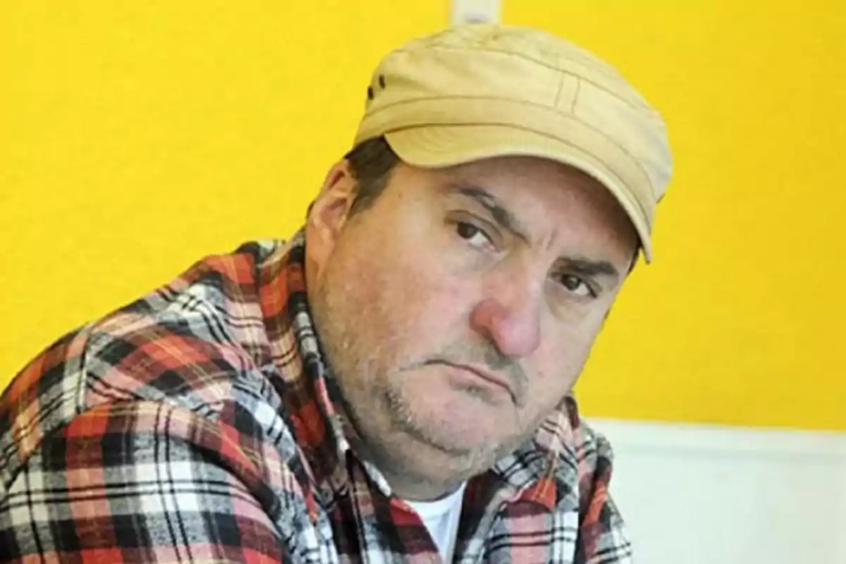Hombre con camisa de cuadros y gorra beige mirando serio frente a un fondo amarillo Hombre con camisa de cuadros y gorra beige mirando serio frente a un fondo amarillo