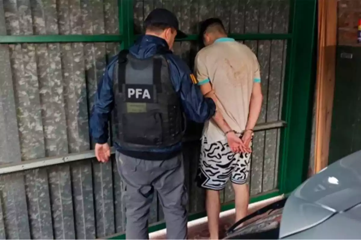 Un policía con chaleco que dice PFA detiene a un hombre esposado frente a una puerta metálica. Un policía con chaleco que dice PFA detiene a un hombre esposado frente a una puerta metálica.