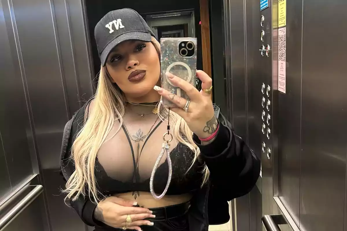 Blonde-haired woman wearing a black cap taking a selfie in an elevator while wearing a sheer black top and a jacket