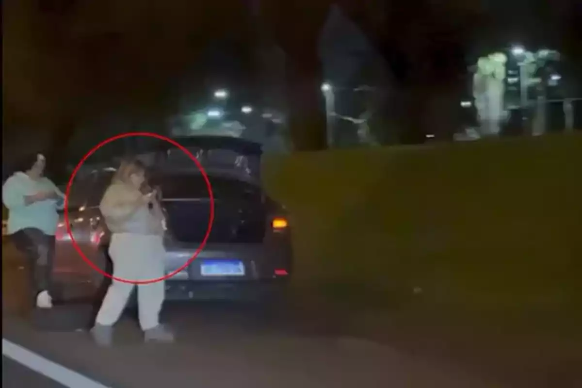 Two women are next to a car stopped on the road at night, one of them is inside a red circle and the trunk of the vehicle is open. Two women are next to a car stopped on the road at night, one of them is inside a red circle and the trunk of the vehicle is open.