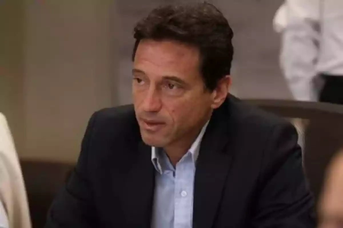 Dark-haired man wearing a navy blue blazer and light blue jersey sitting at a table in an office setting