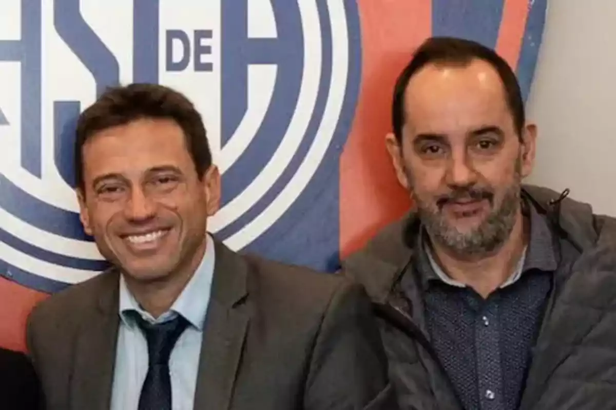 Two men pose together in front of a backdrop with a sports club emblem; one of them is wearing a suit and tie while the other is wearing a casual jacket. Two men pose together in front of a backdrop with a sports club emblem; one of them is wearing a suit and tie while the other is wearing a casual jacket.