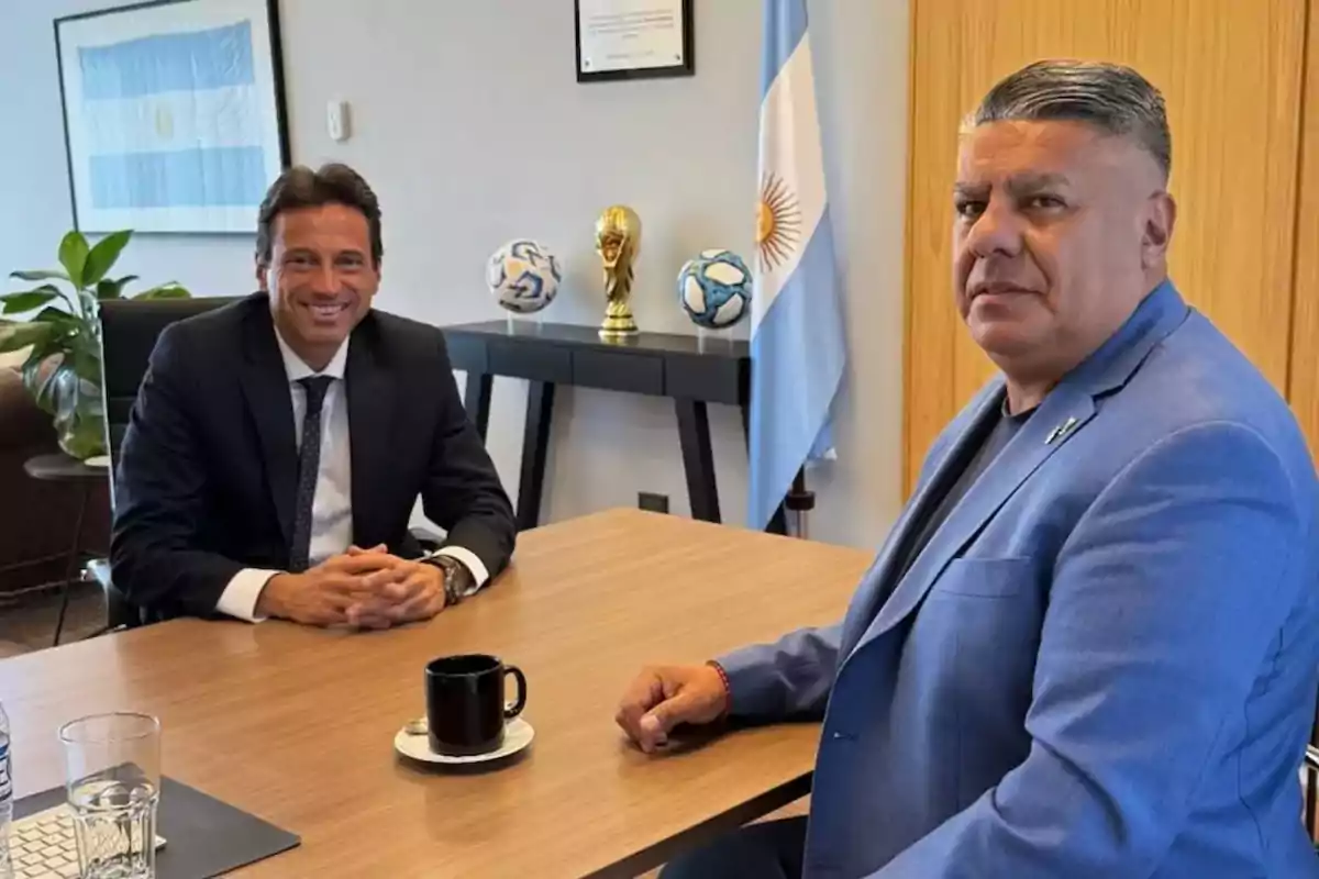 Two men in suits sitting in an office with an Argentine flag, a plant, a table with cups, and a replica of the World Cup in the background.