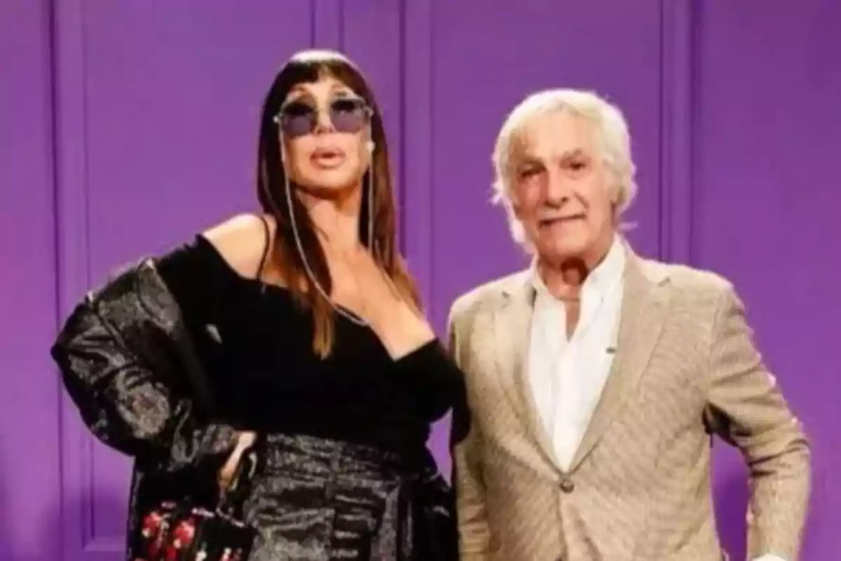 Two people pose in front of a violet wall, a woman wearing sunglasses and a gray-haired man in a light jacket.