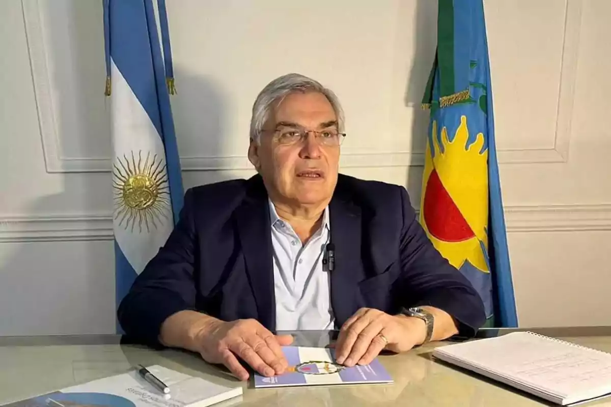 Hombre de cabello canoso y gafas sentado en un escritorio con dos banderas detrás, vistiendo saco azul y camisa clara, con libros y una libreta sobre la mesa. Hombre de cabello canoso y gafas sentado en un escritorio con dos banderas detrás, vistiendo saco azul y camisa clara, con libros y una libreta sobre la mesa.