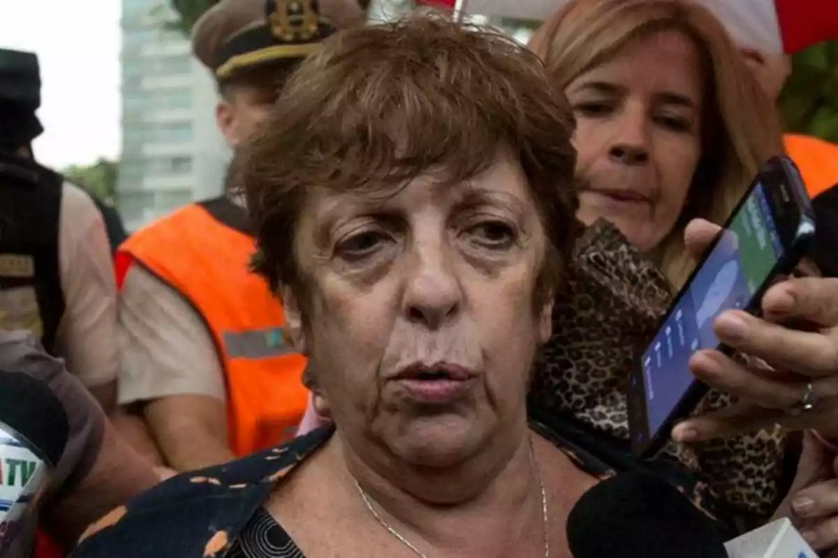 Mujer de cabello corto y castaño hablando frente a varios micrófonos rodeada de periodistas y personas con chalecos naranjas