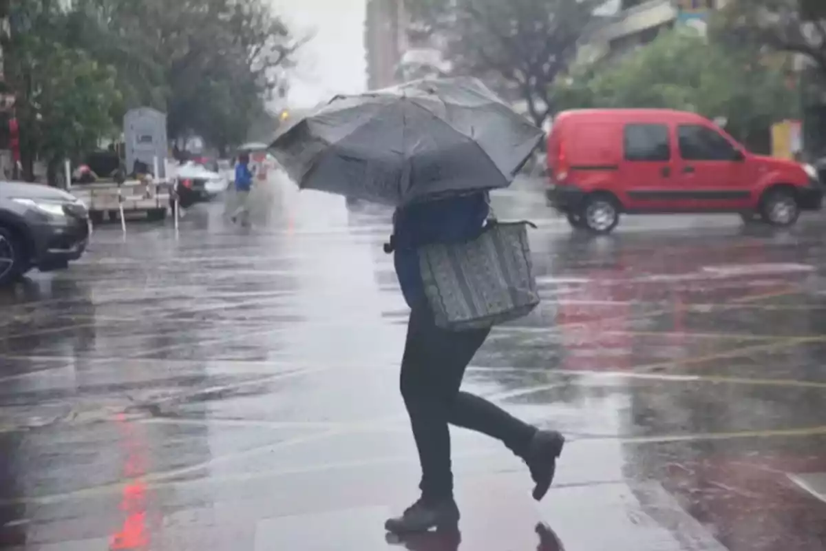 Person crossing a street in the rain with a black umbrella and a large bag while the pavement is wet and cars can be seen in the background Person crossing a street in the rain with a black umbrella and a large bag while the pavement is wet and cars can be seen in the background