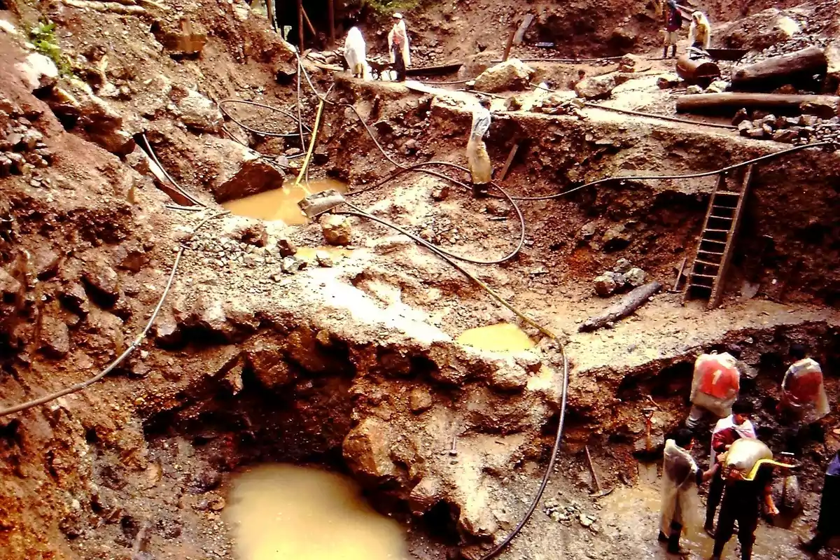 Personas trabajando en una mina artesanal de tierra y lodo con charcos de agua y herramientas manuales