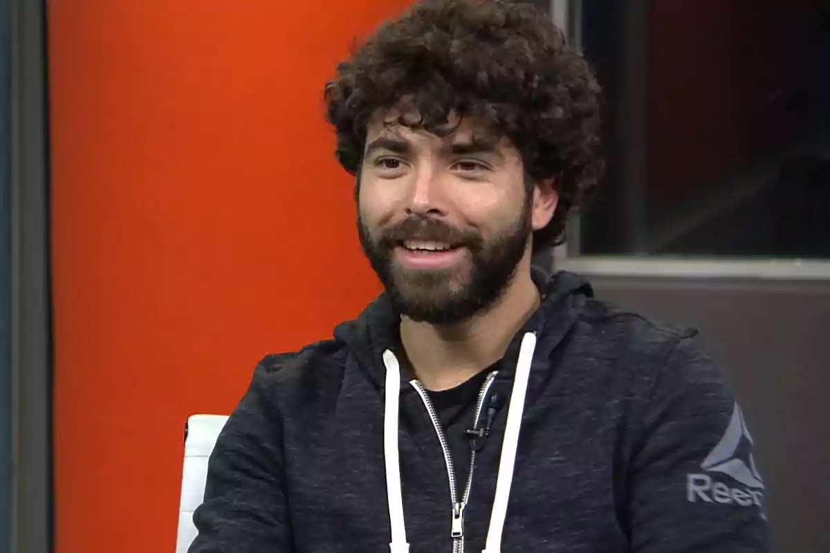 Hombre joven de cabello rizado y barba, sonriendo mientras está sentado con una sudadera oscura frente a un fondo naranja Hombre joven de cabello rizado y barba, sonriendo mientras está sentado con una sudadera oscura frente a un fondo naranja