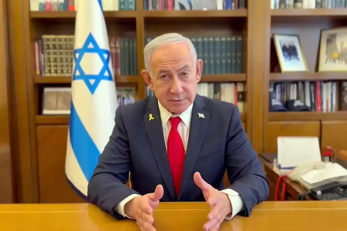 Hombre de cabello canoso con traje oscuro y corbata roja sentado en un escritorio con una bandera de Israel y estanterías de libros al fondo