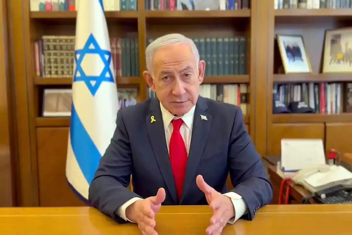 Gray-haired man in a dark suit and red tie sitting at a desk with an Israeli flag and a bookshelf in the background Gray-haired man in a dark suit and red tie sitting at a desk with an Israeli flag and a bookshelf in the background