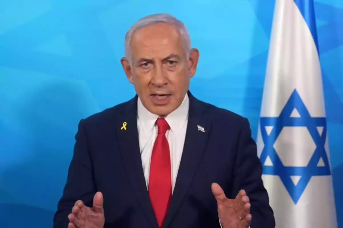 Gray-haired man in a dark suit and red tie speaking in front of an Israeli flag against a blue background Gray-haired man in a dark suit and red tie speaking in front of an Israeli flag against a blue background