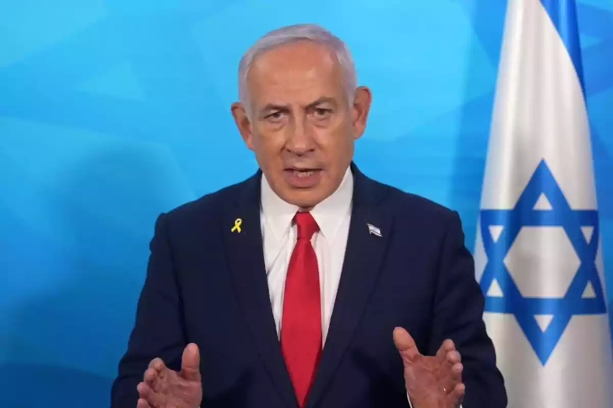 Gray-haired man in a dark suit and red tie speaking in front of an Israeli flag and a blue background Gray-haired man in a dark suit and red tie speaking in front of an Israeli flag and a blue background