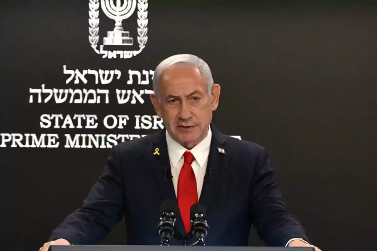 Gray-haired man in a dark suit and red tie speaking in front of microphones with a black background and Hebrew and English letters.