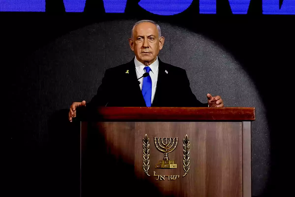 A man in a dark suit and blue tie is standing behind a wooden podium with the national emblem of Israel as he gives a speech.