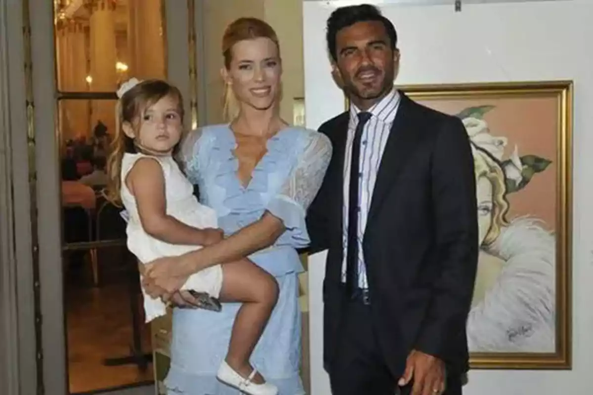Una mujer con un vestido celeste sostiene a una niña pequeña mientras un hombre con traje oscuro está a su lado y detrás de ellos hay un cuadro enmarcado