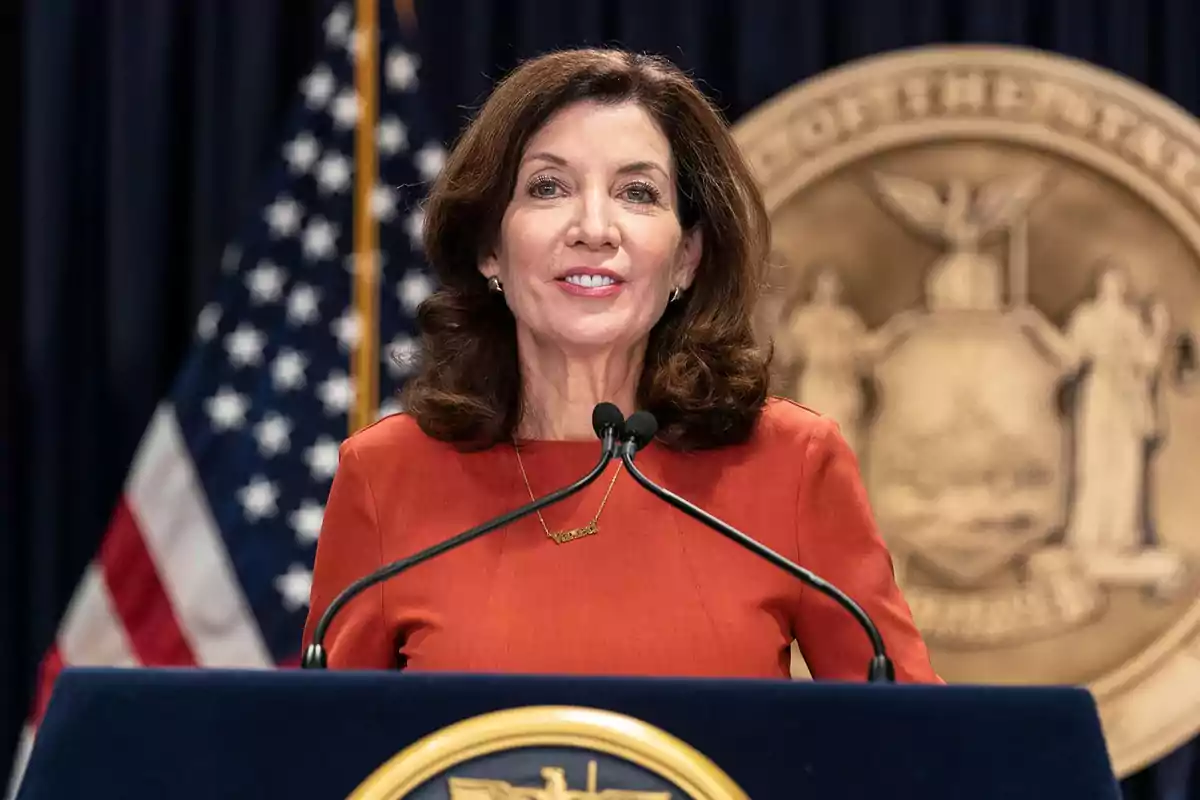 Brown-haired woman in an orange dress speaking at a podium with microphones, with a United States flag and a golden emblem in the background