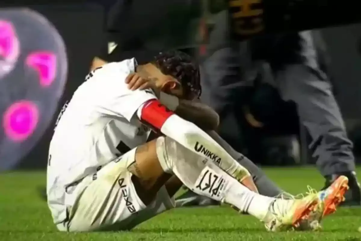 Futbolista sentado en el césped con el rostro cubierto por los brazos mostrando una expresión de tristeza