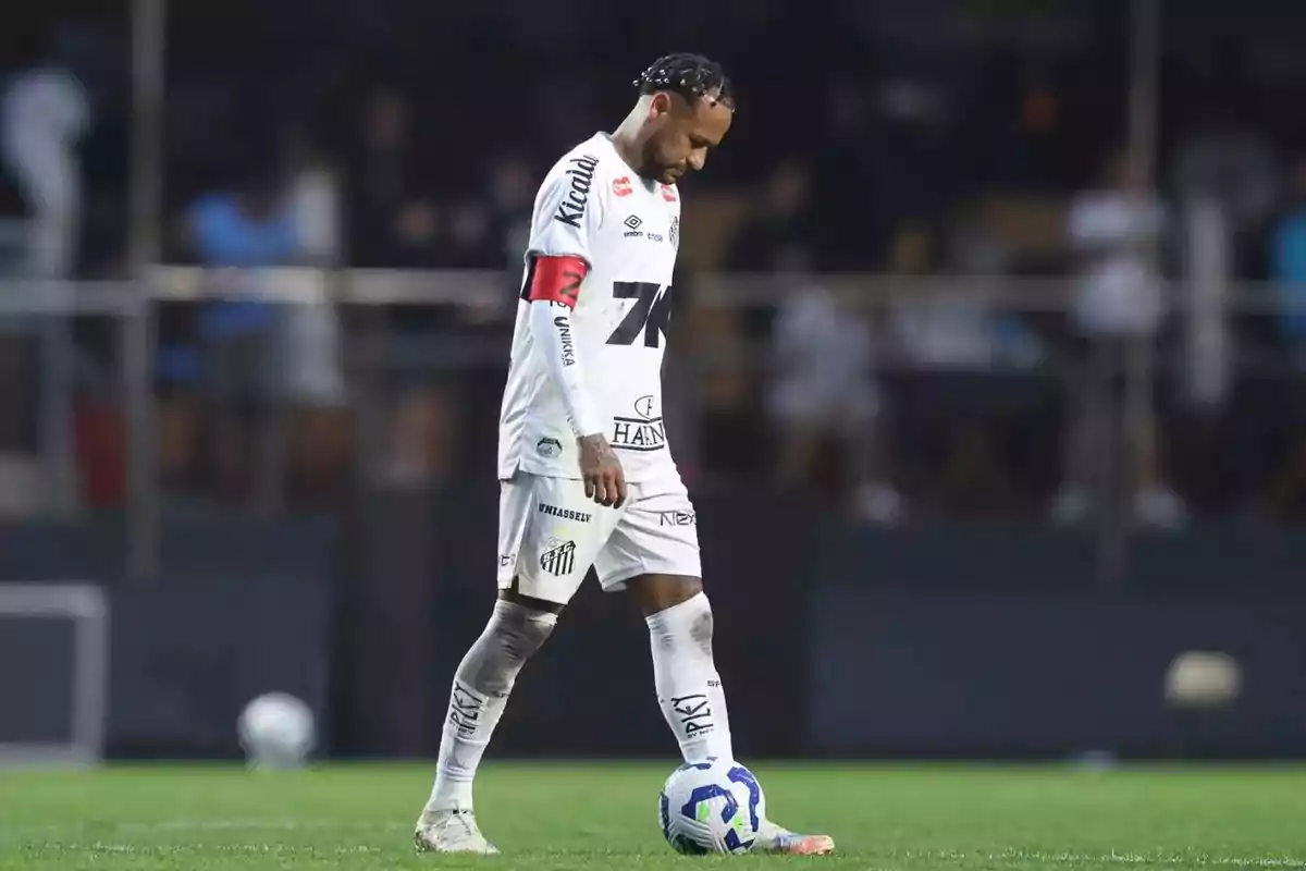 Futbolista con uniforme blanco y cinta de capitán roja mira hacia abajo mientras está de pie junto a un balón en el campo Futbolista con uniforme blanco y cinta de capitán roja mira hacia abajo mientras está de pie junto a un balón en el campo